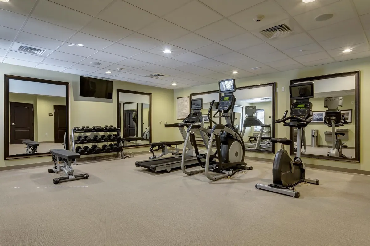 The fitness center is equipped with modern exercise machines, a wall of mirrors, dumbbells, and a television monitor. The energetic space invites guests to maintain their routines or try group fitness activities, embodying the convenience of 24/7 amenities.