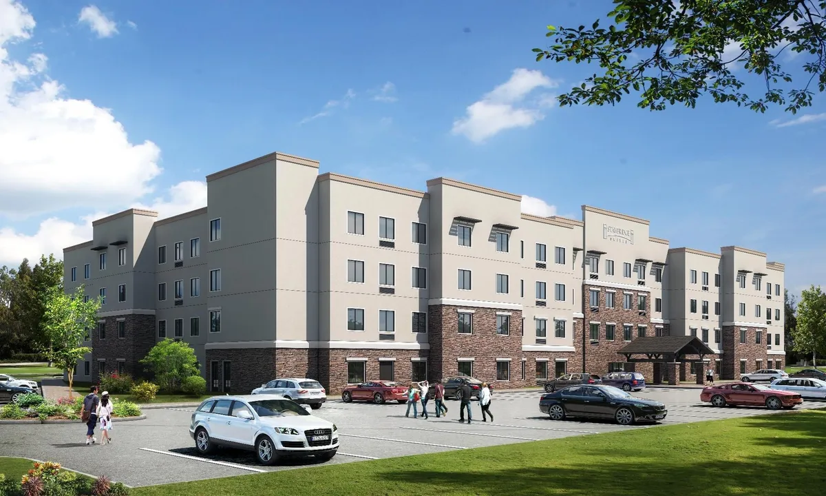 A wide exterior view of Staybridge Suites St Louis - Westport, featuring its modern building facade, a welcoming entrance awning, well-maintained landscaping, and a clearly visible parking lot. The environment appears inviting, showing guests arriving or socializing near the entrance, with the hotel visually integrated into its neighborhood.