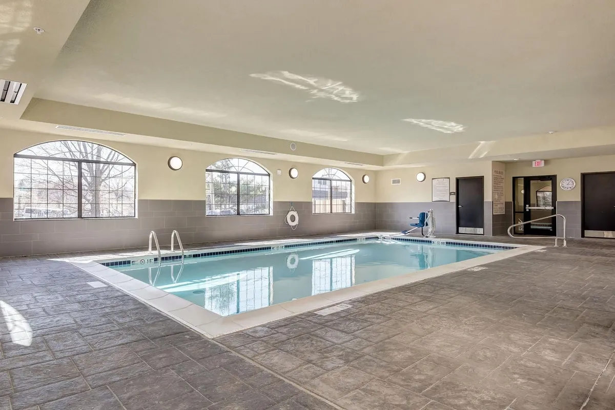 This image features a bright indoor swimming pool with large windows allowing plenty of natural light, comfortable poolside seating, and visible safety equipment. The tranquil setting is ideal for both swimming and relaxation, reinforcing the sense of a hotel-style retreat.