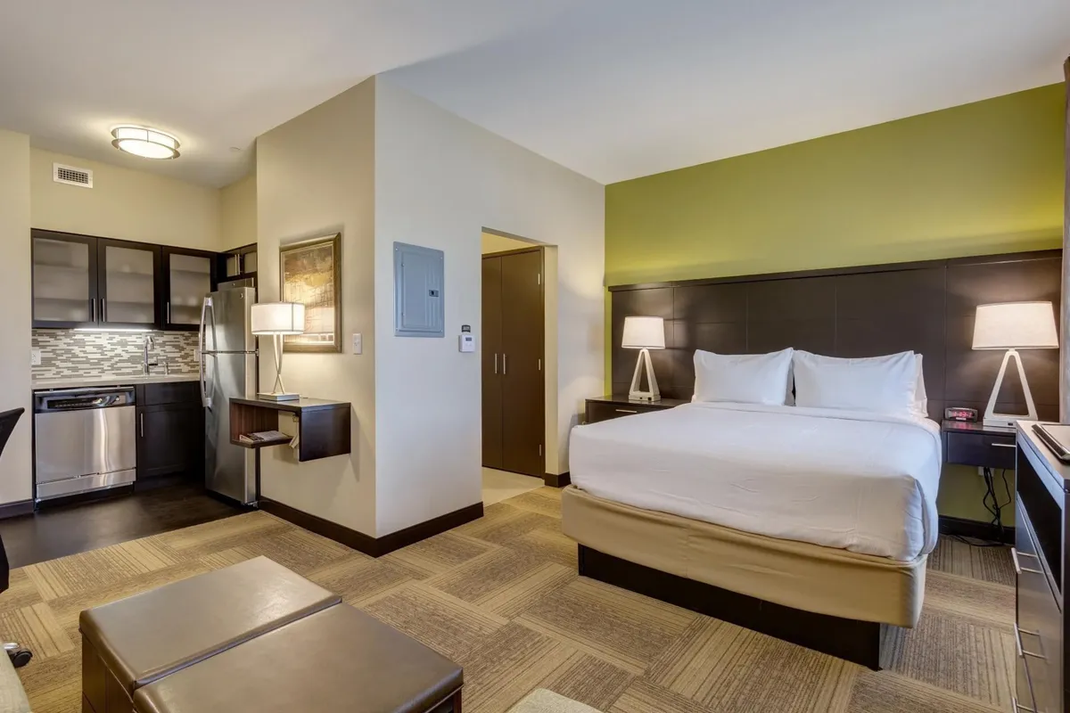 The image showcases a bright, inviting studio with a plush bed dressed in crisp white linens, a functional kitchenette perfect for preparing meals, warm bedside lighting, and tasteful artwork that adds a homey touch. The overall atmosphere is cozy and practical, ideal for both relaxation and productivity.
