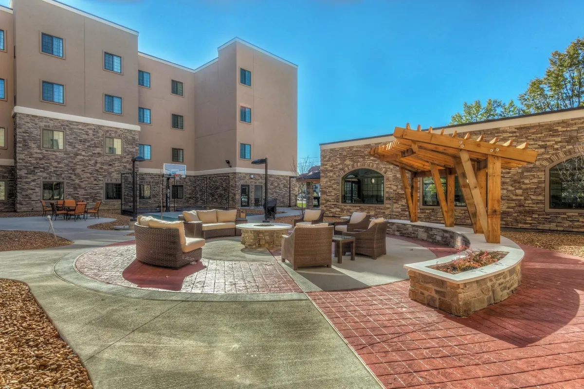 An inviting outdoor seating area with a fire pit beneath a pergola, surrounded by lush landscaping. The space is designed for relaxation, informal gatherings, or unwinding after work, suggesting both leisure and community amid a business trip.