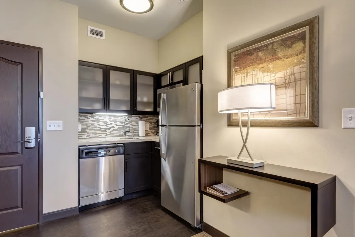 A modern space with stainless steel appliances, contemporary cabinetry, decorative wall art, and a table lamp. The arrangement suggests a multi-functional living area, blending style, practicality, and a touch of personality in its décor.