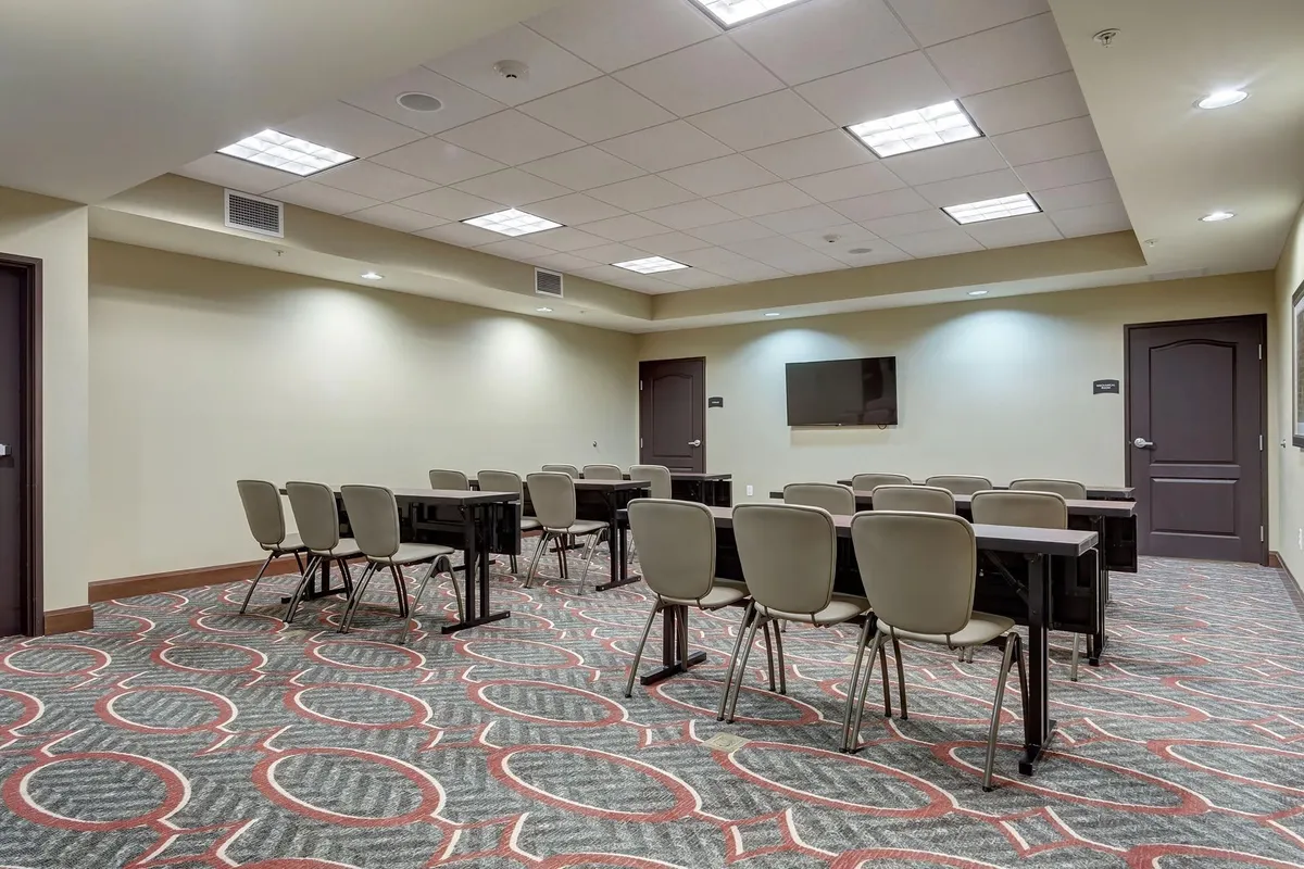The image shows a well-lit common area with tables and chairs arranged for group gatherings. A television and patterned carpet add to the inviting, functional feel. The space is suitable for meetings, quiet work, or gathering with family, offering both privacy and a communal atmosphere.
