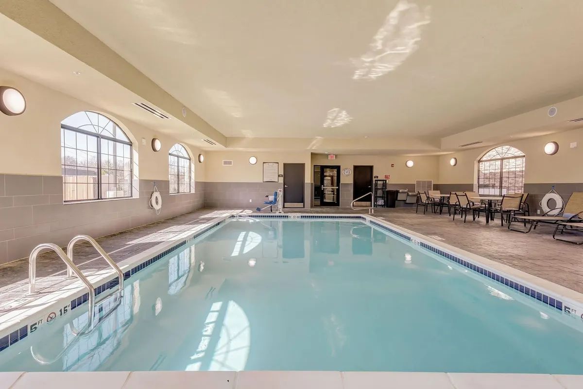This image features an inviting indoor swimming pool area, with large windows letting in natural light. Poolside seating provides a comfortable place to relax, while modern lighting adds to the tranquil atmosphere. The scene conveys a sense of calm, comfort, and the possibility for quiet moments of relaxation or gentle activity.